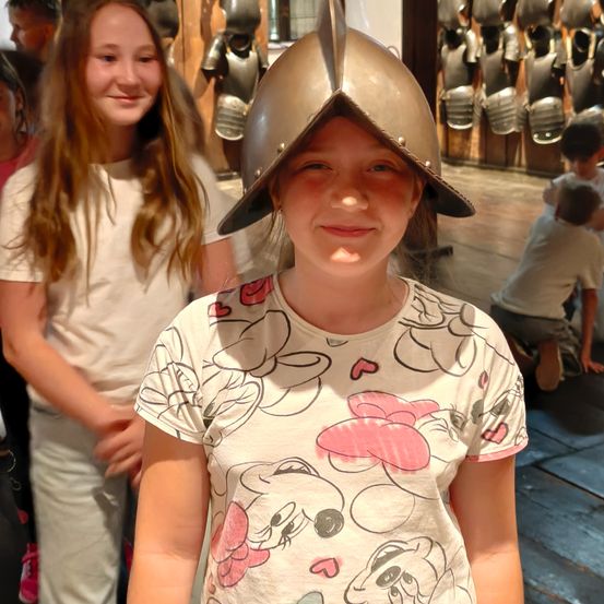 Ein junges Mädchen mit einem silbernen Helm mit Visier steht vor einer Wand mit Rüstungsausstellung. Ein weiteres Mädchen steht hinter ihr und ein Junge sitzt auf dem Boden im Hintergrund.