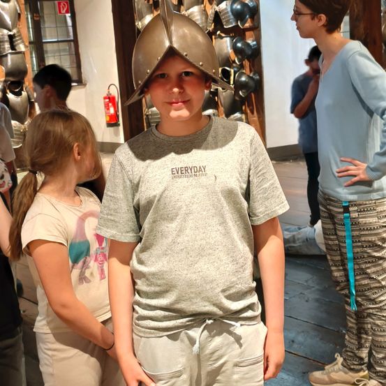 Ein junger Junge mit einem mittelalterlichen Helm steht vor einer Ausstellung von Rüstungen und Waffen. Hinter ihm schauen ein Mädchen und eine Frau auf die Ausstellung. Im Hintergrund steht ein Mann in der Nähe eines Feuerlöschers.