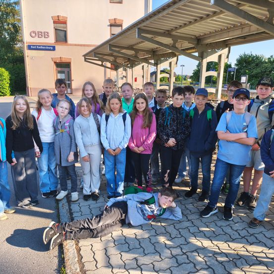 Eine Gruppe von Kindern und Jugendlichen posiert für ein Foto an einer Bushaltestelle vor einem Gebäude mit der Aufschrift 'Bad Radkersburg'.