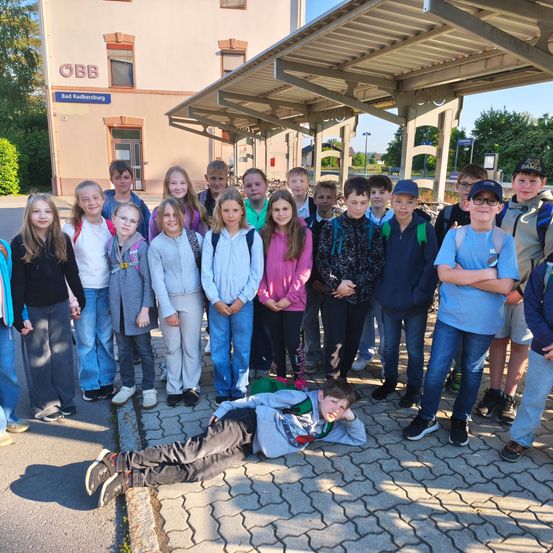 Eine Gruppe von Kindern und Jugendlichen steht und sitzt auf einem Bürgersteig vor einem Gebäude mit dem Schild 'Bad Radkersburg.' Einige tragen Rucksäcke und Freizeitkleidung.