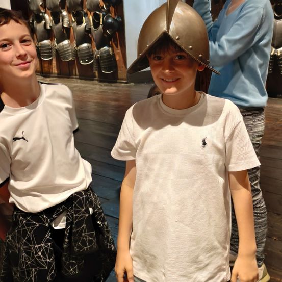 Zwei junge Jungen stehen vor einer Wand mit mittelalterlicher Rüstung. Ein Junge trägt einen Helm, während der andere lächelt. Eine Frau in einem blauen Shirt steht hinter ihnen.