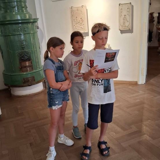Drei Kinder stehen in einem Raum mit Holzböden, eines liest ein Papier, während ein grüner Heizkörper und Kunstwerke an den Wänden hängen.