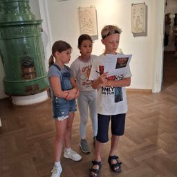 Drei Kinder stehen in einem Raum mit Holzböden, eines liest ein Papier, während ein grüner Heizkörper und Kunstwerke an den Wänden hängen.