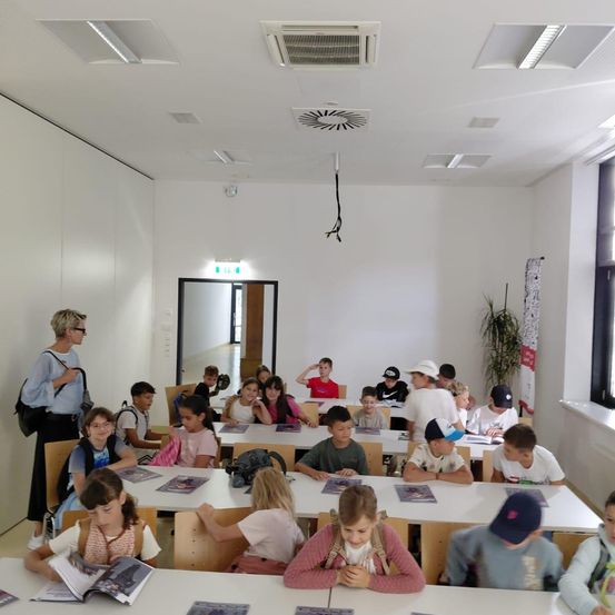 Ein Klassenzimmer mit Schülern, die an Tischen sitzen, Bücher lesen und eine Frau ansehen, die in der Nähe der Wand steht.
