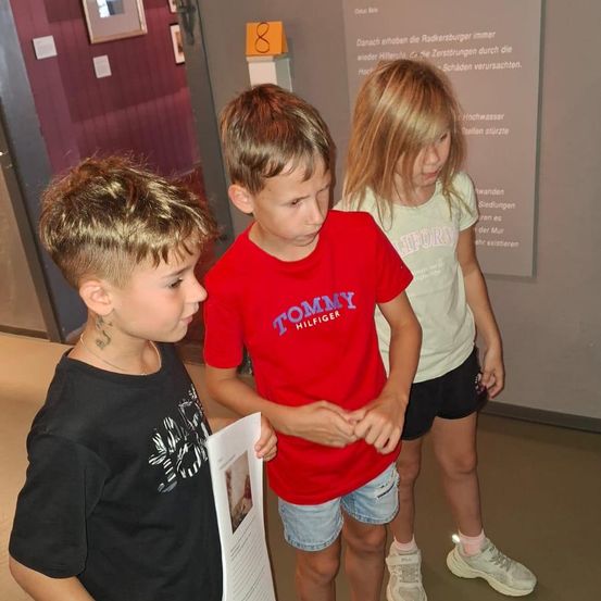 Drei Kinder, zwei Jungen und ein Mädchen, stehen in einem Museum und betrachten ein Stück Papier. Der Junge in der Mitte trägt ein rotes Tommy Hilfiger-Shirt.