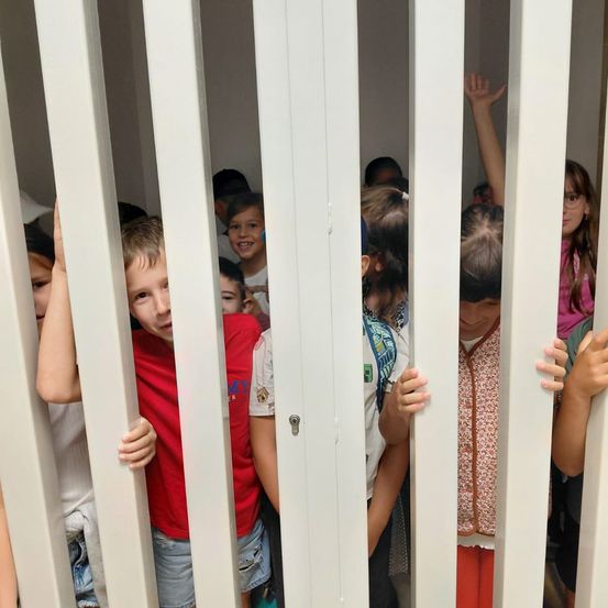 Eine Gruppe von Kindern lugt durch die Lücken zwischen weißen Türen, einige lächeln und andere sehen neugierig aus.