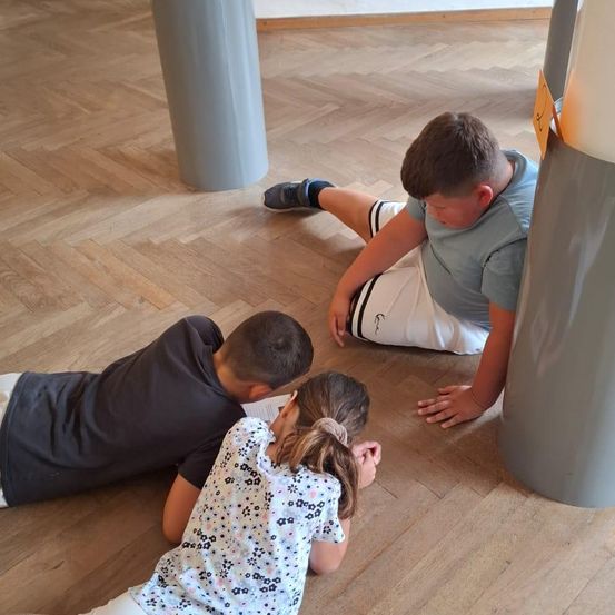 Drei Kinder liegen auf einem Holzboden und lesen. Ein Junge liegt auf dem Rücken, ein Mädchen auf der Seite und ein anderer Junge sitzt.