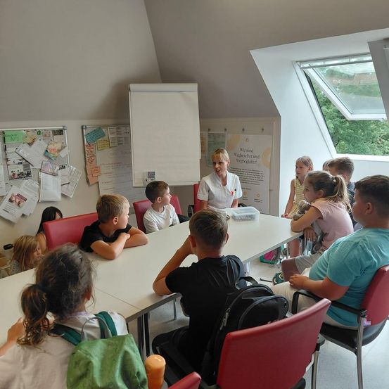 Eine Gruppe von Kindern und einer Frau in einer weißen Jacke sitzen um einen Tisch in einem Klassenzimmer. Papiere sind an der Wand hinter ihnen angeheftet.