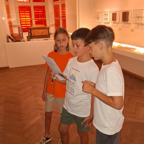 Drei Kinder stehen in einem Museum und schauen auf ein Stück Papier. Der Junge auf der linken Seite hält einen Stift. Hinter ihnen ist eine Vitrine mit verschiedenen Gegenständen zu sehen.