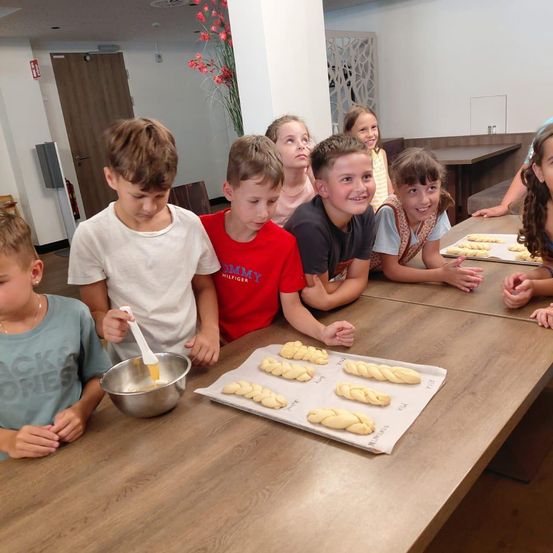 Eine Gruppe von Kindern versammelt sich um einen Tisch und bereitet gebratenes Brot zu. Einige kneten Teig in einer Schüssel, während andere zuschauen. Tabletts mit gebratenem Teig liegen auf dem Tisch.