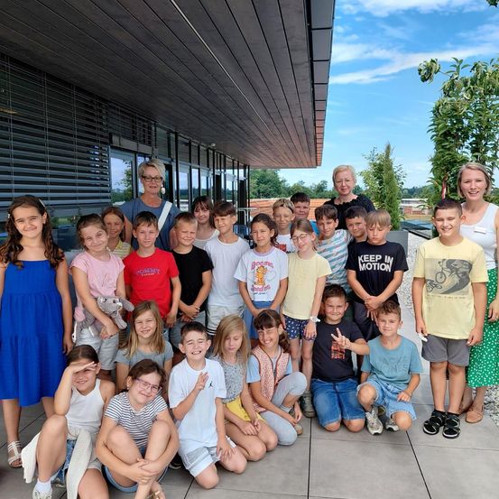 Eine Gruppe von Kindern und Erwachsenen, darunter drei Frauen, posiert für ein Foto vor einem Gebäude. Die Kinder tragen Freizeitkleidung, und einige halten Kuscheltiere. Die Frauen stehen hinten.