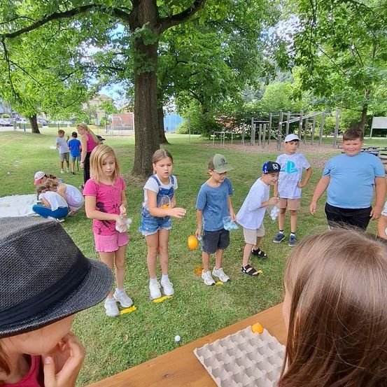Kinder spielen ein Spiel in einem Park mit einem Rasenfeld, Bäumen und einem Holztisch mit Eiern in einer Schale. Einige tragen Mützen und Turnschuhe.