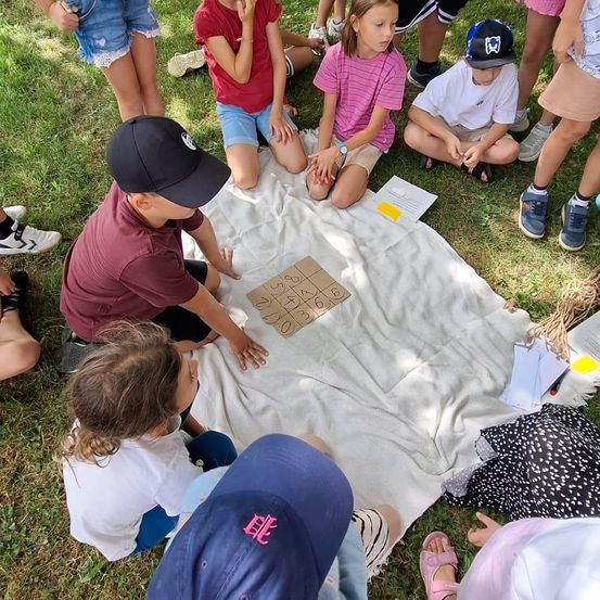 Eine Gruppe von Kindern sitzt auf einer Decke im Gras, einige spielen ein Brettspiel.