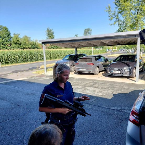 Eine Polizistin in Uniform steht in einem Parkplatz, hält ein Gewehr und spricht mit einem Kind. Hinter ihr sind drei Autos unter einem Carport geparkt.