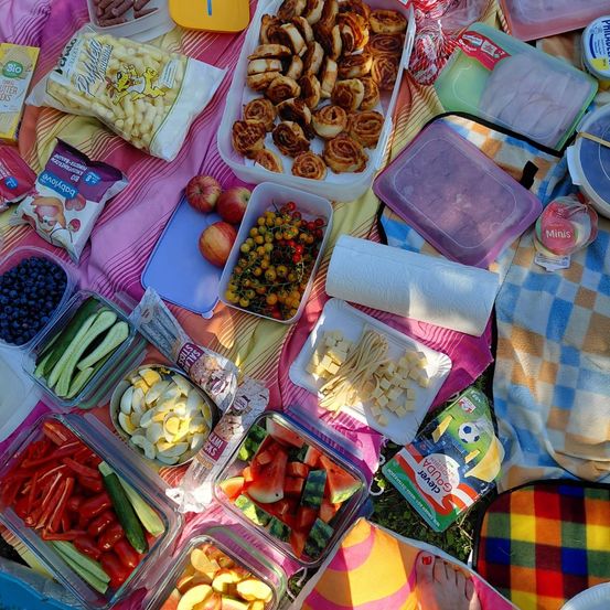 Ein Picknick-Arrangement mit verschiedenen Lebensmitteln wie Obst, Gemüse, Gebäck und Snacks auf einer bunten Decke.