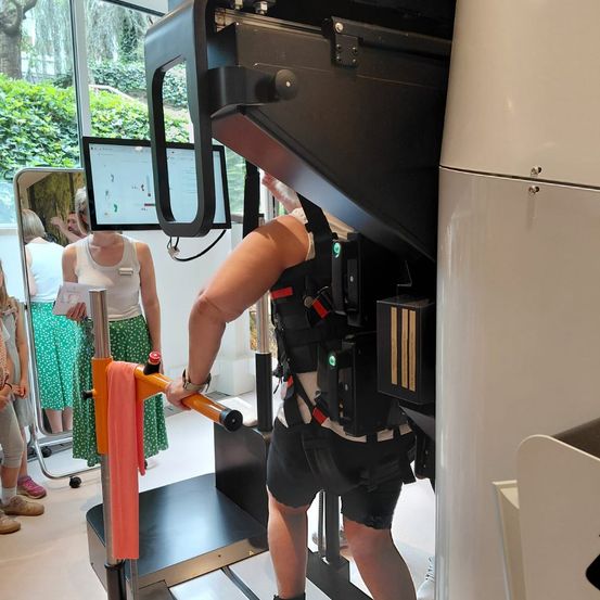 Ein Mann mit einem Gurt und Shorts benutzt ein Laufband mit einem Monitor, der ein Diagramm anzeigt. Hinter ihm steht eine Frau mit einem grünen Rock.