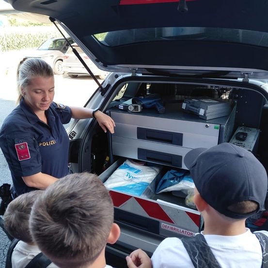 Eine Polizistin zeigt Kindern den Inhalt des Kofferraums eines Fahrzeugs, einschließlich eines Defibrillators, medizinischer Vorräte und Ausrüstung.