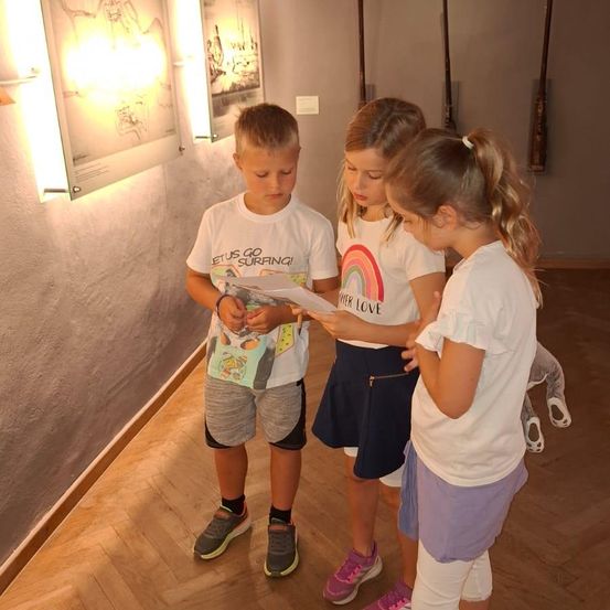 Drei Kinder, zwei Mädchen und ein Junge, stehen in einem Flur und lesen ein Blatt Papier. Der Junge trägt ein weißes T-Shirt mit den Worten 'Lasst uns surfen gehen!' Die Mädchen tragen weiße T-Shirts mit einem Regenbogen und den Worten 'Niemals Liebe.' Der Flur hat Holzböden und Wände.