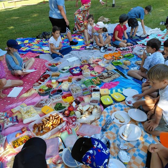 Eine Gruppe von Kindern und Erwachsenen macht ein Picknick auf einer Grasfläche. Sie sitzen um einen Tisch, der mit einem blau-weiß karierten Tuch bedeckt ist. Auf dem Tisch befinden sich verschiedene Essensartikel, darunter Sandwiches, Obst und Gemüse. Einige der Kinder tragen Mützen und Turnschuhe. In der Ferne steht eine Person in der Nähe eines Pfahls.