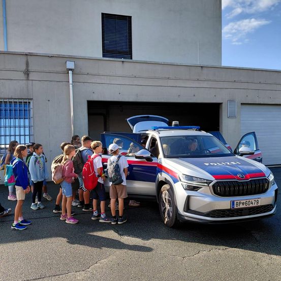 Eine Gruppe von Kindern in Schuluniformen steht vor einem Polizeiauto mit offenen Türen, möglicherweise während eines Schulbesuchs.