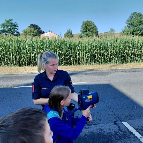 Eine Polizistin steht neben einem kleinen Mädchen, das ein Radargerät in der Hand hält, an einem sonnigen Tag neben einem Maisfeld.