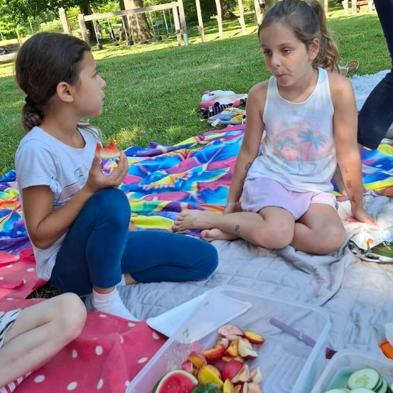 Zwei junge Mädchen sitzen auf einer Decke in einem Park, eine hält ein Stück Wassermelone und die andere schaut sie an. Sie sind von verschiedenen Obst- und Gemüsesorten umgeben.