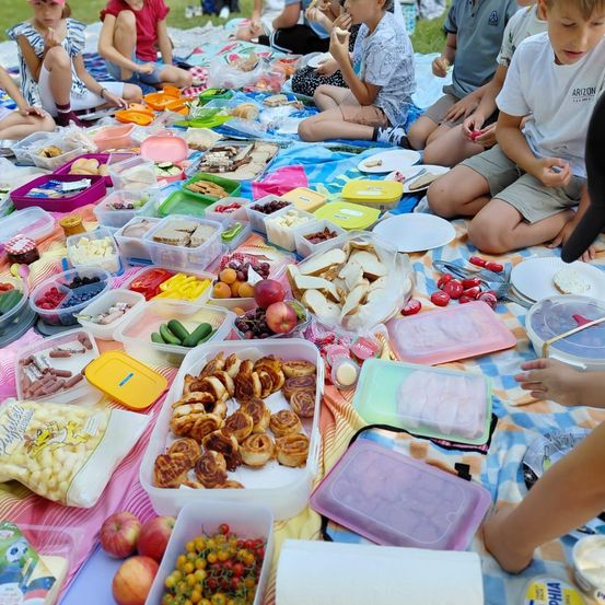 Eine Gruppe von Kindern sitzt auf einer Picknickdecke und isst verschiedene Lebensmittel aus Plastikbehältern und Tellern. Der Tisch ist mit einer bunten Decke bedeckt und hat eine Vielzahl von Obst, Gemüse und anderen Snacks.