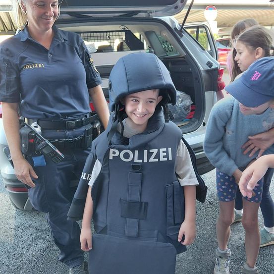 Ein junger Junge, der eine Polizeiweste und einen Helm trägt, steht neben einer Polizeibeamtin mit lächelndem Gesicht. Hinter ihnen schaut ein Mädchen mit einer blauen Mütze den Jungen an.