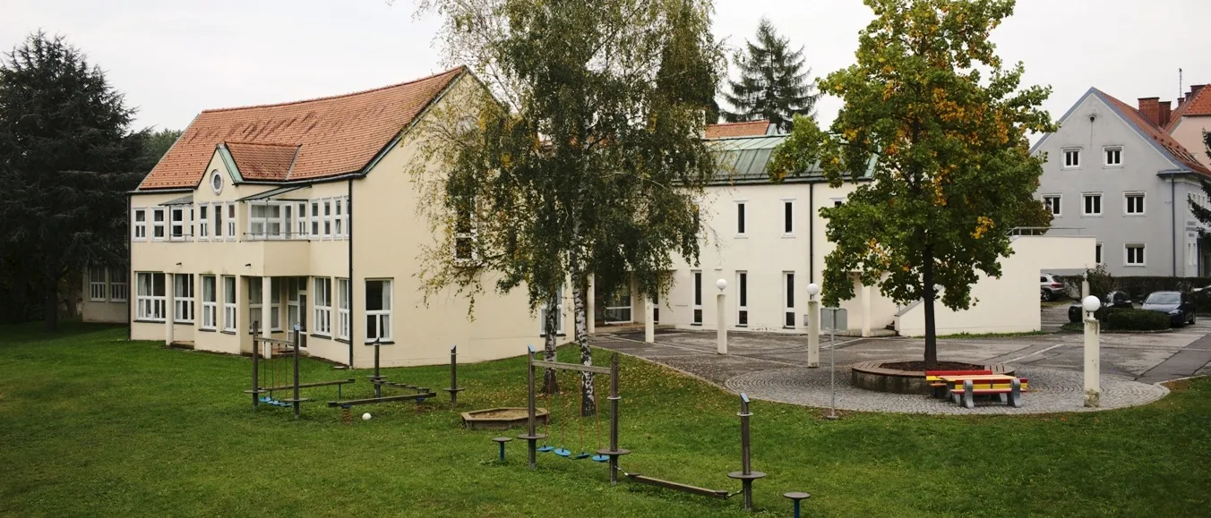 Außenansicht eines Schulgebäudes mit einem braunen Dach, mehreren Fenstern und einem Spielplatz mit Schaukeln und Bänken. Das Gebäude ist von grünem Gras und Bäumen umgeben.