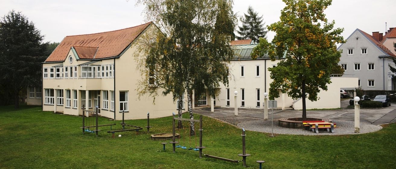 Außenansicht eines Schulgebäudes mit einem braunen Dach, mehreren Fenstern und einem Spielplatz mit Schaukeln und Bänken. Das Gebäude ist von grünem Gras und Bäumen umgeben.