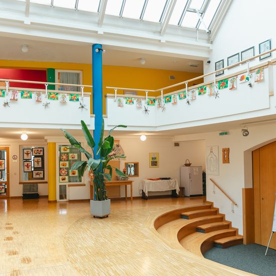 Ein Innenbereich mit Holzboden, Glasdecke und blauem Pfosten. Kunstwerke hängen an der Wand und es gibt eine Topfpflanze auf dem Boden.