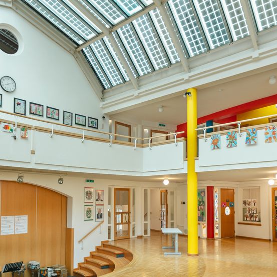 Ein Innenraum eines mehrstöckigen Gebäudes mit Holzböden, einem Glasdach und einer gelben Säule. Bilderrahmen und eine Uhr schmücken die Wände. Treppen führen zu einer unteren Ebene mit Türen und einem offenen Raum.