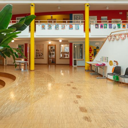 Ein leerer Flur in einem modernen Gebäude mit gelben Säulen, Holzböden und verschiedenen Kunstwerken an den Wänden. Rechts stehen zwei Tische und Stühle.