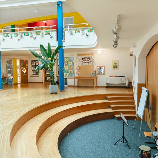Ein Innenauditorium mit einer Holzbühne, Teppichboden und Musikinstrumenten. Ein Topf mit einer Pflanze und eine Tafel stehen auf der Bühne. Der Raum hat Holzwände und Decken.