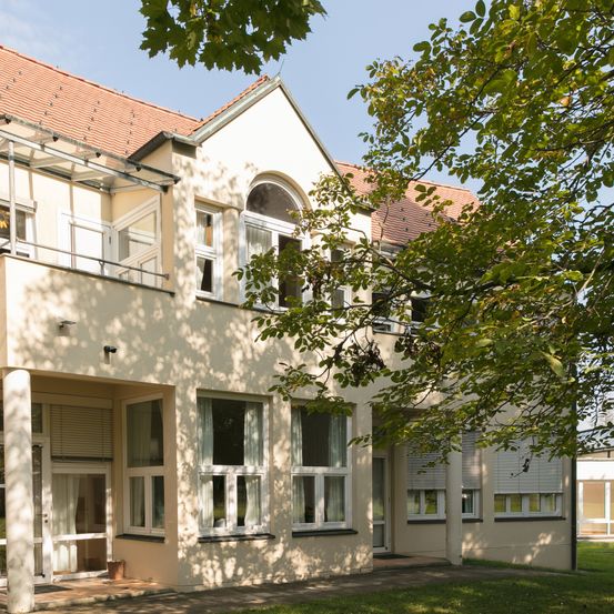 Ein beigefarbenes Gebäude mit rotem Dach und weißen Fenstern unter einem Baum an einem sonnigen Tag.