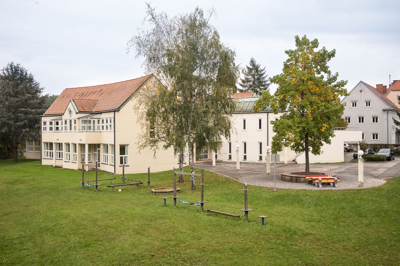Ein großes Gebäude mit einem roten Dach steht in einem Grasbereich. Es hat viele Fenster und einen runden Sitzbereich mit Bänken. Es gibt Spielgeräte und Bäume in der Nähe.