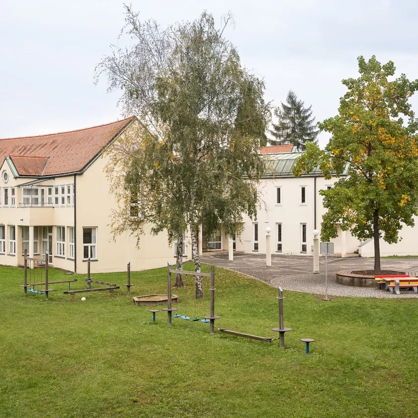 Ein großes Gebäude mit einem roten Dach steht in einem Grasbereich. Es hat viele Fenster und einen runden Sitzbereich mit Bänken. Es gibt Spielgeräte und Bäume in der Nähe.