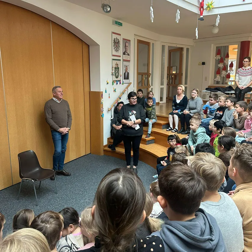 Eine Gruppe von Kindern sitzt auf einer Holztreppe und hört einem Mann zu, der vor ihnen steht. Hinter ihnen sitzt eine Frau auf den Stufen. Der Raum hat eine Holzwand und Decke mit hängenden Dekorationen.