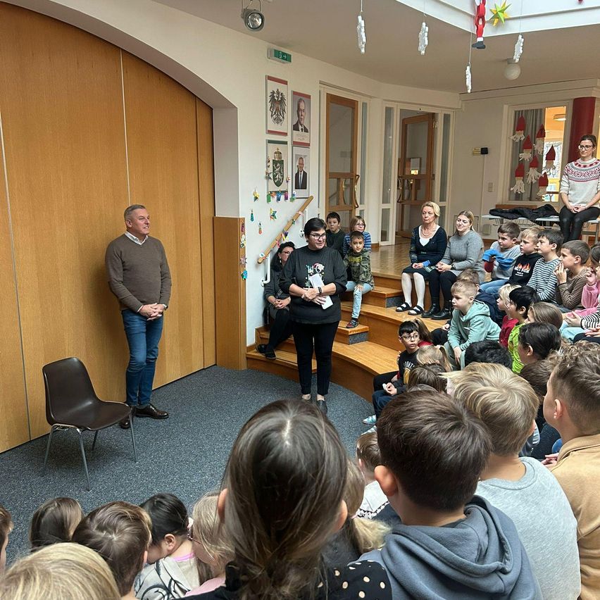 Eine Gruppe von Kindern sitzt auf einer Holztreppe und hört einem Mann zu, der vor ihnen steht. Hinter ihnen sitzt eine Frau auf den Stufen. Der Raum hat eine Holzwand und Decke mit hängenden Dekorationen.