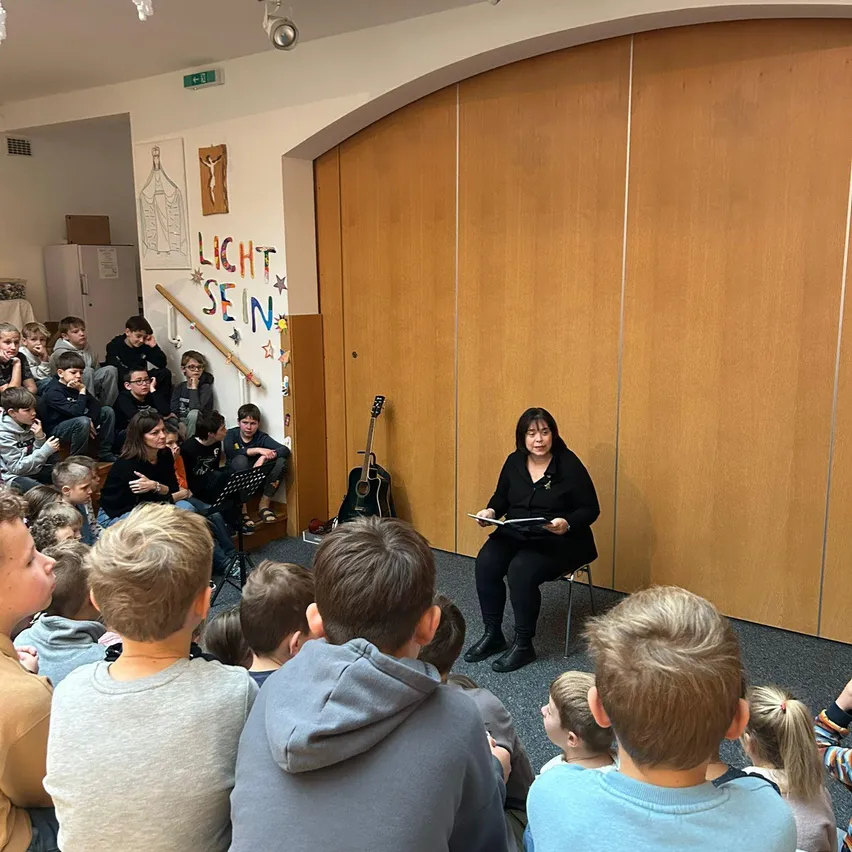 Eine Frau liest vor einer Gruppe von Kindern, die in einem Kreis sitzen, mit einer hölzernen Wand und einem Schild, das 'Licht sein' dahinter steht.