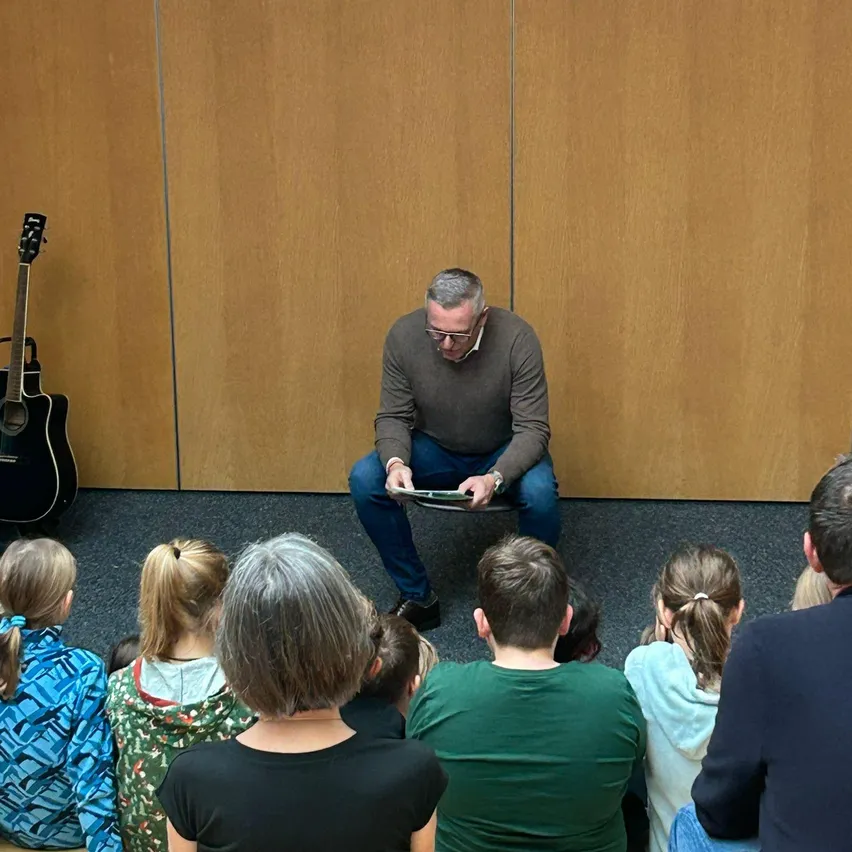 Ein Mann liest Kindern und Erwachsenen, die in einem Kreis auf dem Boden sitzen, ein Buch vor. Eine Gitarre ist gegen eine Holzwand gelehnt.