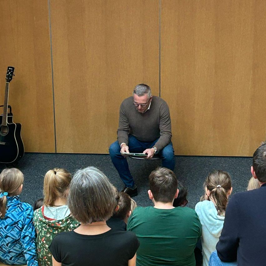 Ein Mann liest Kindern und Erwachsenen, die in einem Kreis auf dem Boden sitzen, ein Buch vor. Eine Gitarre ist gegen eine Holzwand gelehnt.