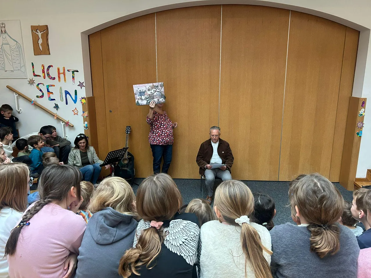 In einem Raum liest ein Mann aus einem Buch, während er sitzt, während eine Frau in der Nähe ein Buch hält. Eine Gitarre steht auf einem Ständer hinter ihr. Ein Publikum aus Kindern sitzt vor ihnen.