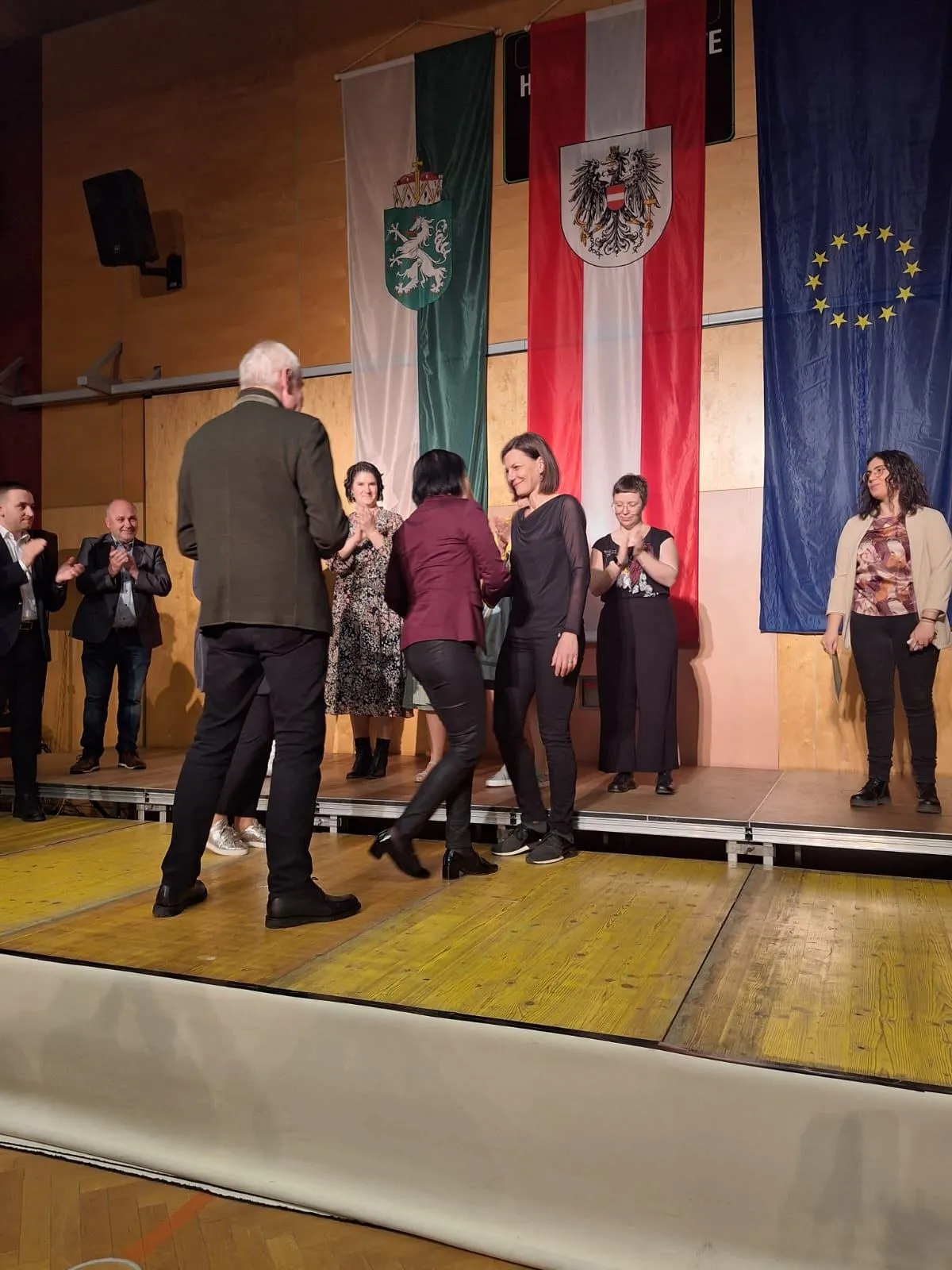 Eine Gruppe von Menschen steht auf einer Bühne, mit Fahnen im Hintergrund. Ein Mann in Anzug klatscht, und drei Frauen stehen in der Mitte.