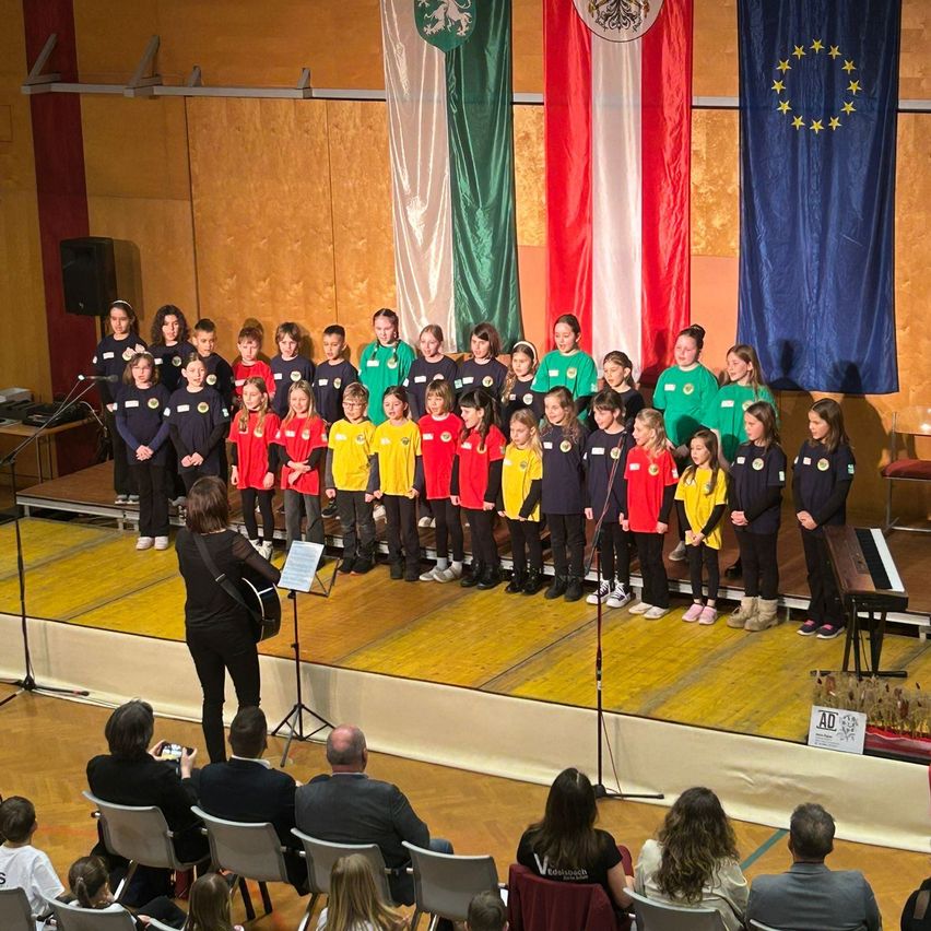 Ein Chor von Kindern in bunten Shirts steht auf der Bühne mit einem Pianisten und Flaggen im Hintergrund. Ein Publikum schaut von Stühlen im Vordergrund zu.