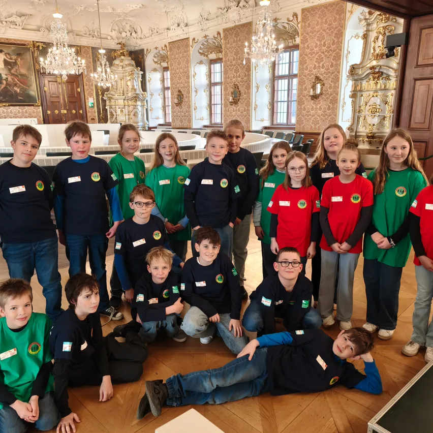 Eine Gruppe junger Kinder in einheitlichen Kleidungsstücken posiert für ein Foto in einem großen Saal mit prunkvoller Einrichtung, Kronleuchtern und Holzböden.