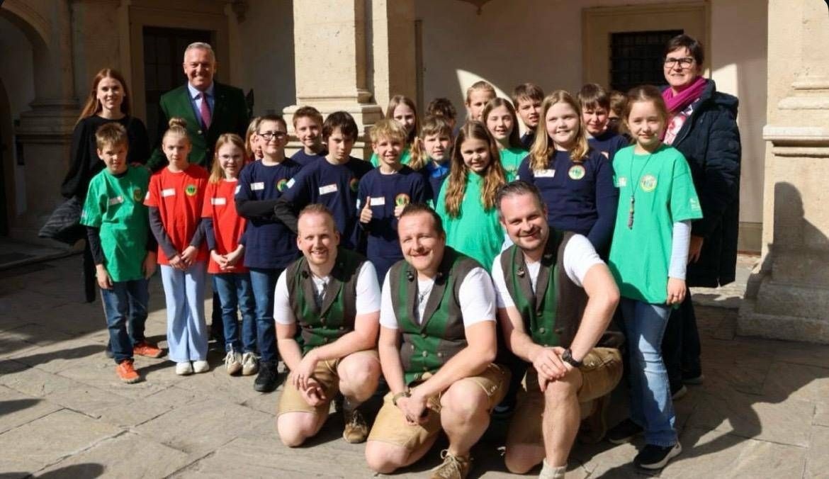 Gruppenfoto von Menschen in grünen und roten Shirts mit einem Mann im Anzug, der vor einem Gebäude lächelt.