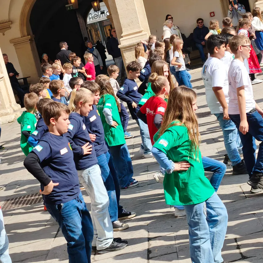 Eine Gruppe von Kindern in einheitlichen Shirts steht und tanzt auf einem öffentlichen Platz. Einige Erwachsene beobachten. Im Hintergrund befindet sich ein Gebäude mit einem gewölbten Eingang.