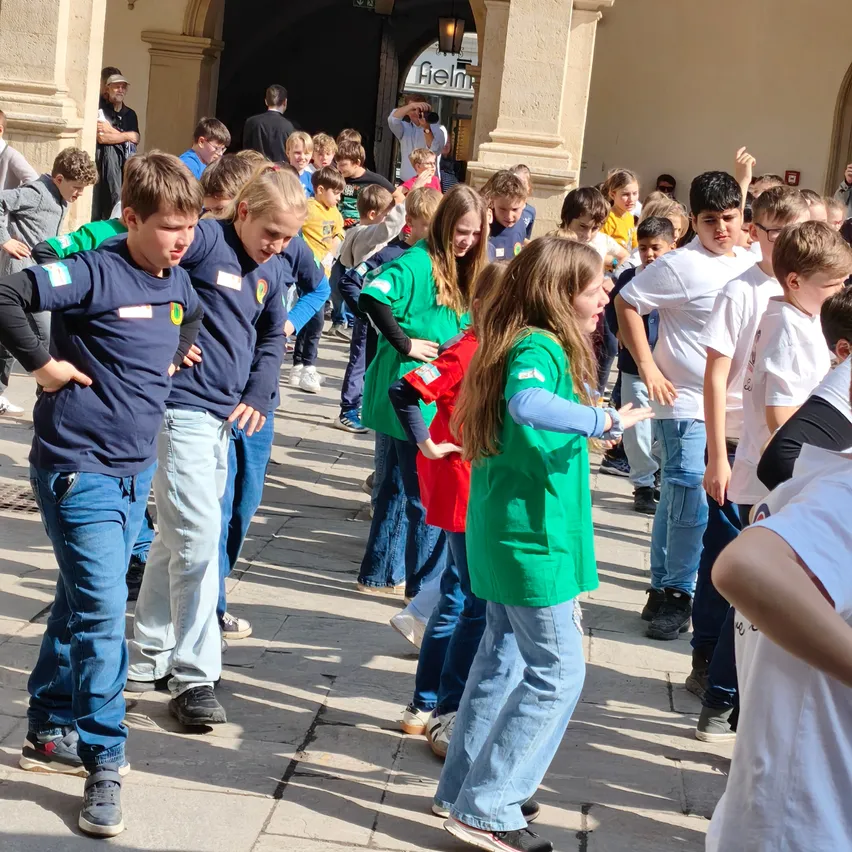 Eine Gruppe von Kindern, einige in blauen und grünen Shirts, tanzt vor einem Gebäude mit einem Torbogen. Ein Mann in einem weißen Shirt beobachtet im Hintergrund.