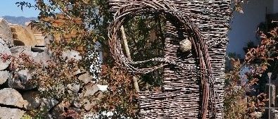 Eine große gewebte Struktur steht in einem Garten mit grünem Gras und braunen Ästen. Sie ist aus Zweigen gefertigt und hat ein Loch in der Mitte. Dahinter befinden sich Pflanzen und eine Steinmauer.